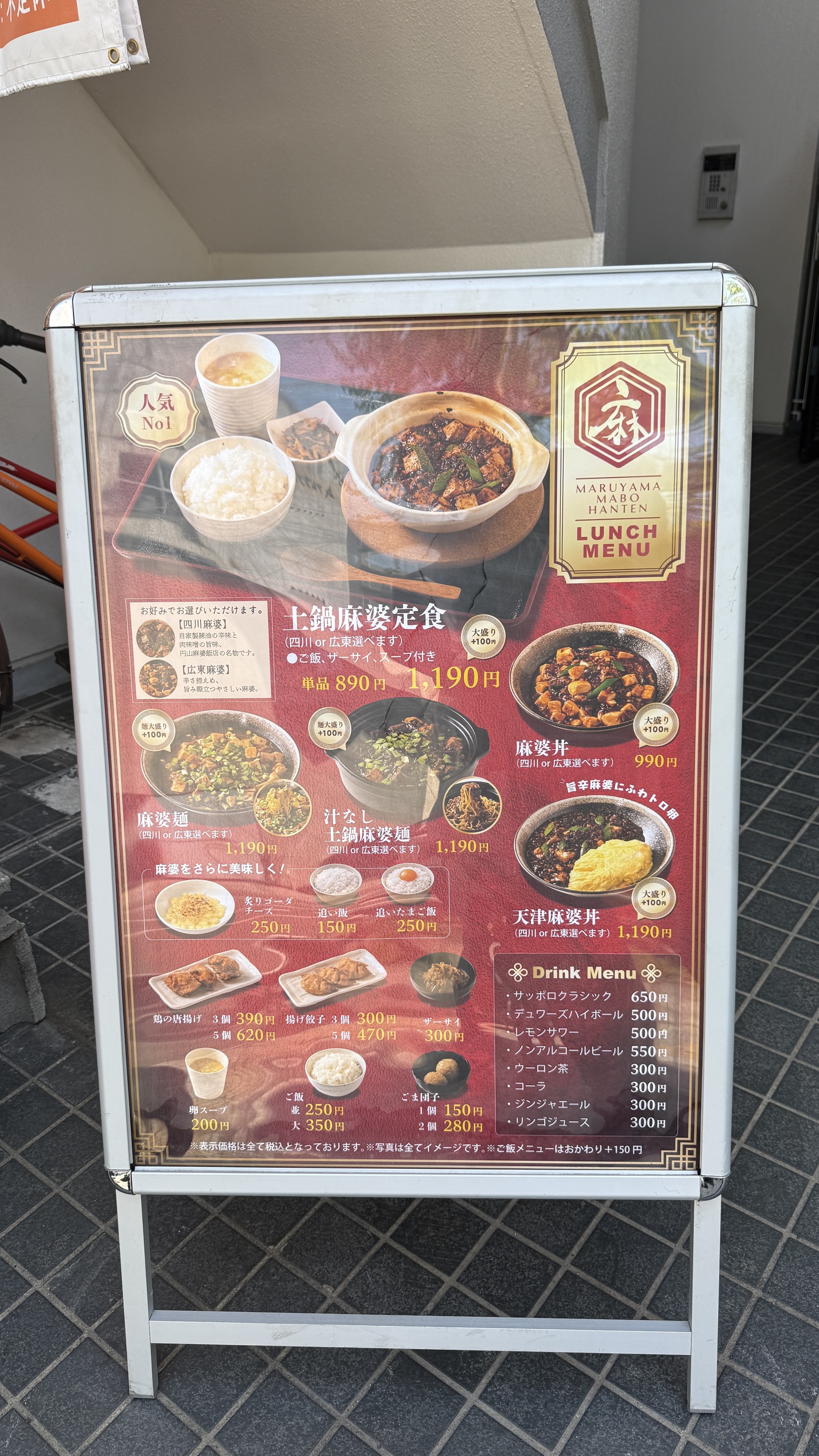 円山麻婆飯店の店外ランチメニュー看板。黒いベースに料理写真が並び、人気No.1の土鍋麻婆定食（単品890円・セット1,190円）が中央に。麻婆丼990円、ハーフ&ハーフの七鍋麻婆定食1,190円、天津麻婆丼1,190円なども。下段にはドリンクメニューも掲載