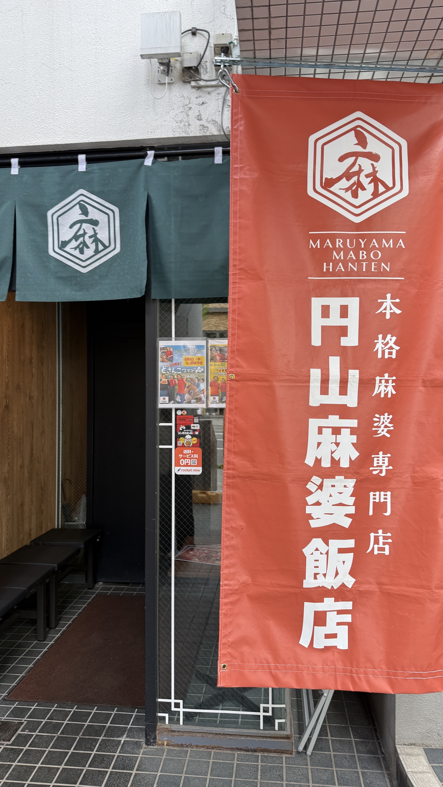 円山麻婆飯店の店舗入口。グレーグリーンの暖簾に円山麻婆飯店のロゴマーク、その横に大きな朱色の幟（のぼり）「MARUYAMA MABO HANTEN 本格麻婆専門店 円山麻婆飯店」。店内に続くウッドフロアと自販機