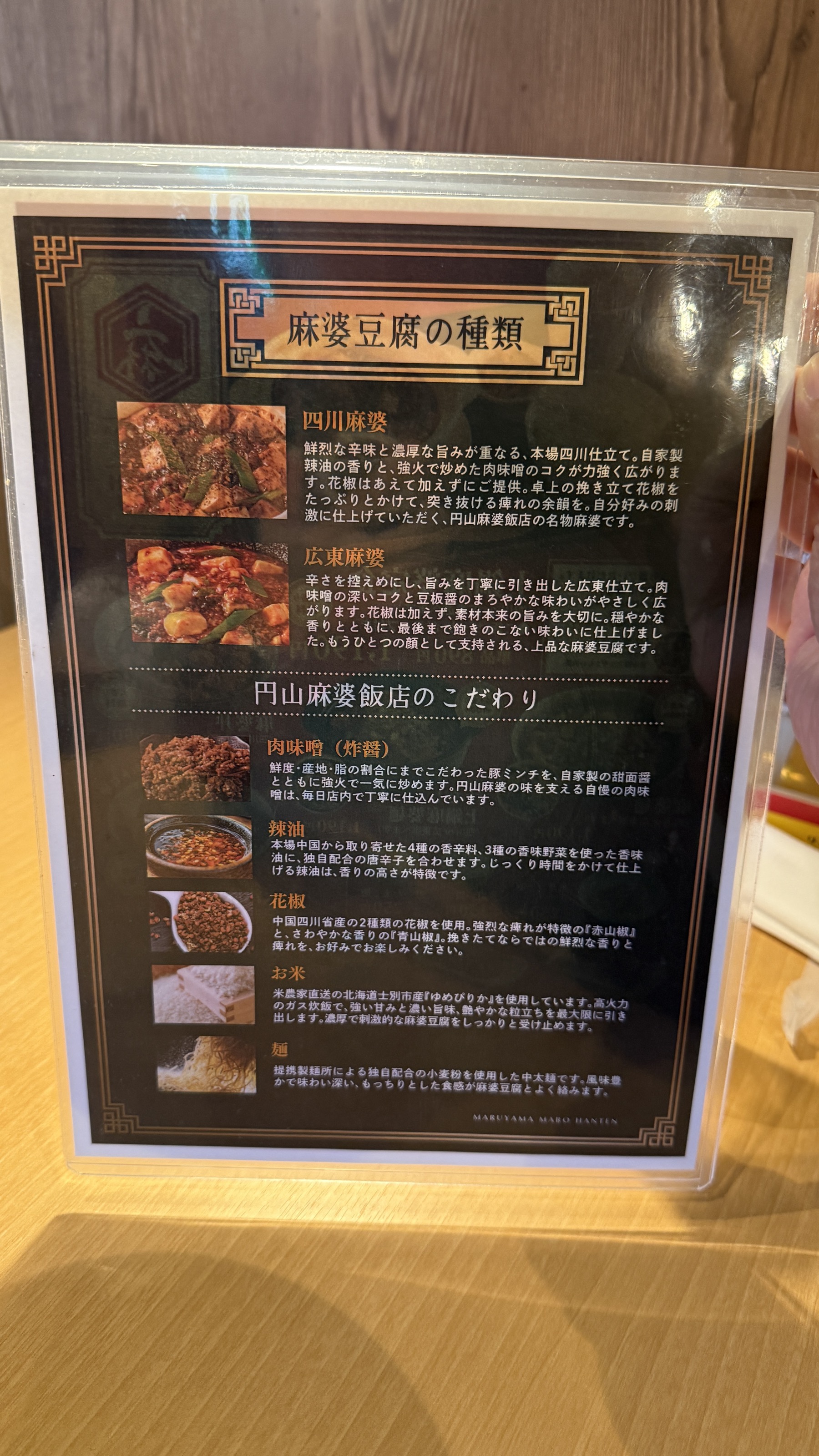 円山麻婆飯店の卓上説明書「麻婆豆腐の種類」。四川麻婆（甜麺醤・豆豉・花椒・ラー油で痺れと香りが強い辛口）と広東麻婆（甘めの味噌ベースでマイルド・辛さ控えめ）の違いが写真付きで説明されている。下段には「円山麻婆飯店のこだわり」として肉味噌・花椒・油・お米・水の解説