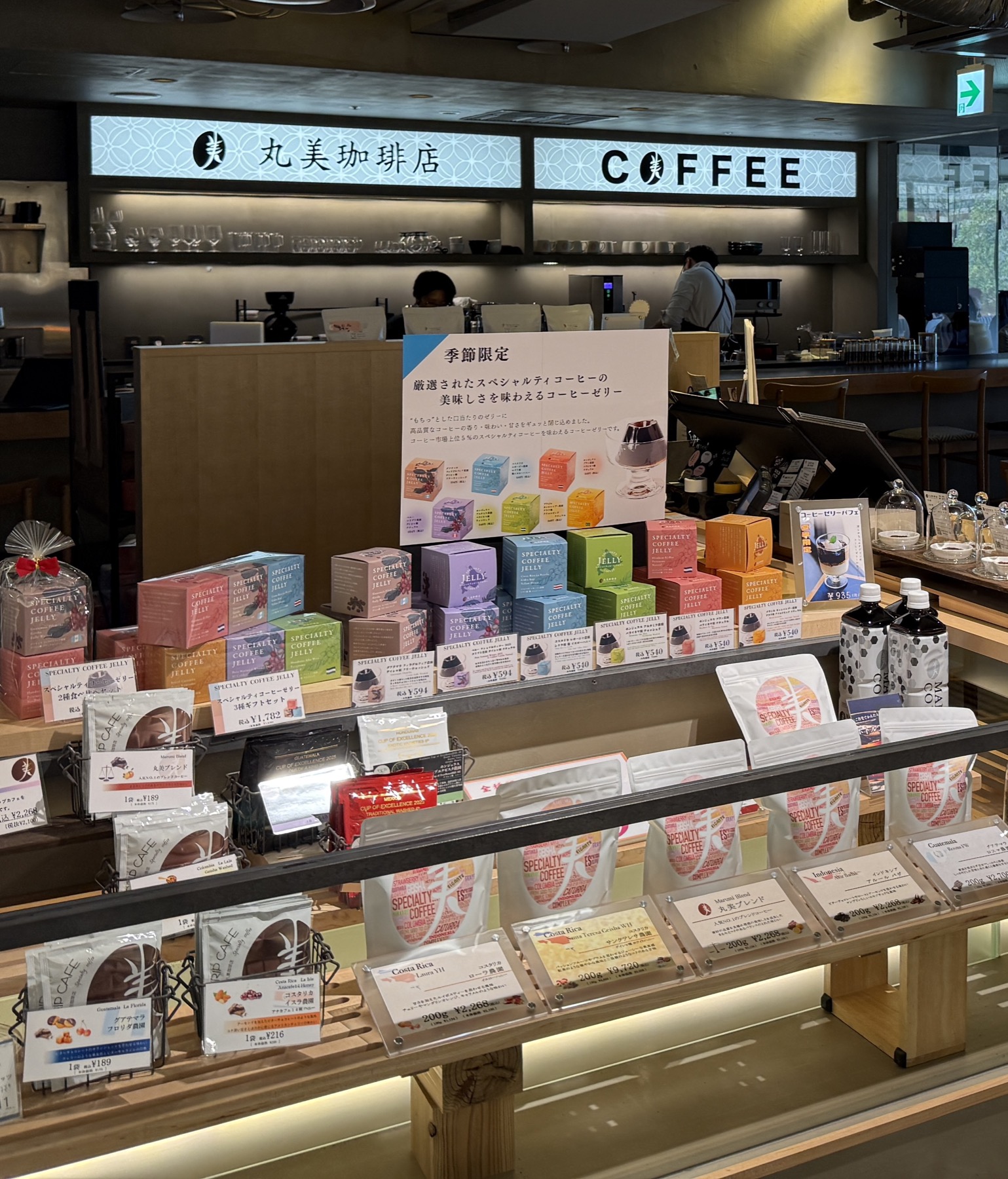丸美珈琲店の店頭。上部に「丸美珈琲店 COFFEE」のロゴ看板、奥にカウンター、手前には季節限定スペシャルティコーヒーゼリーのポップとカラフルなコーヒーゼリーの箱、ドリップバッグなどのギフト商品が並ぶ陳列棚