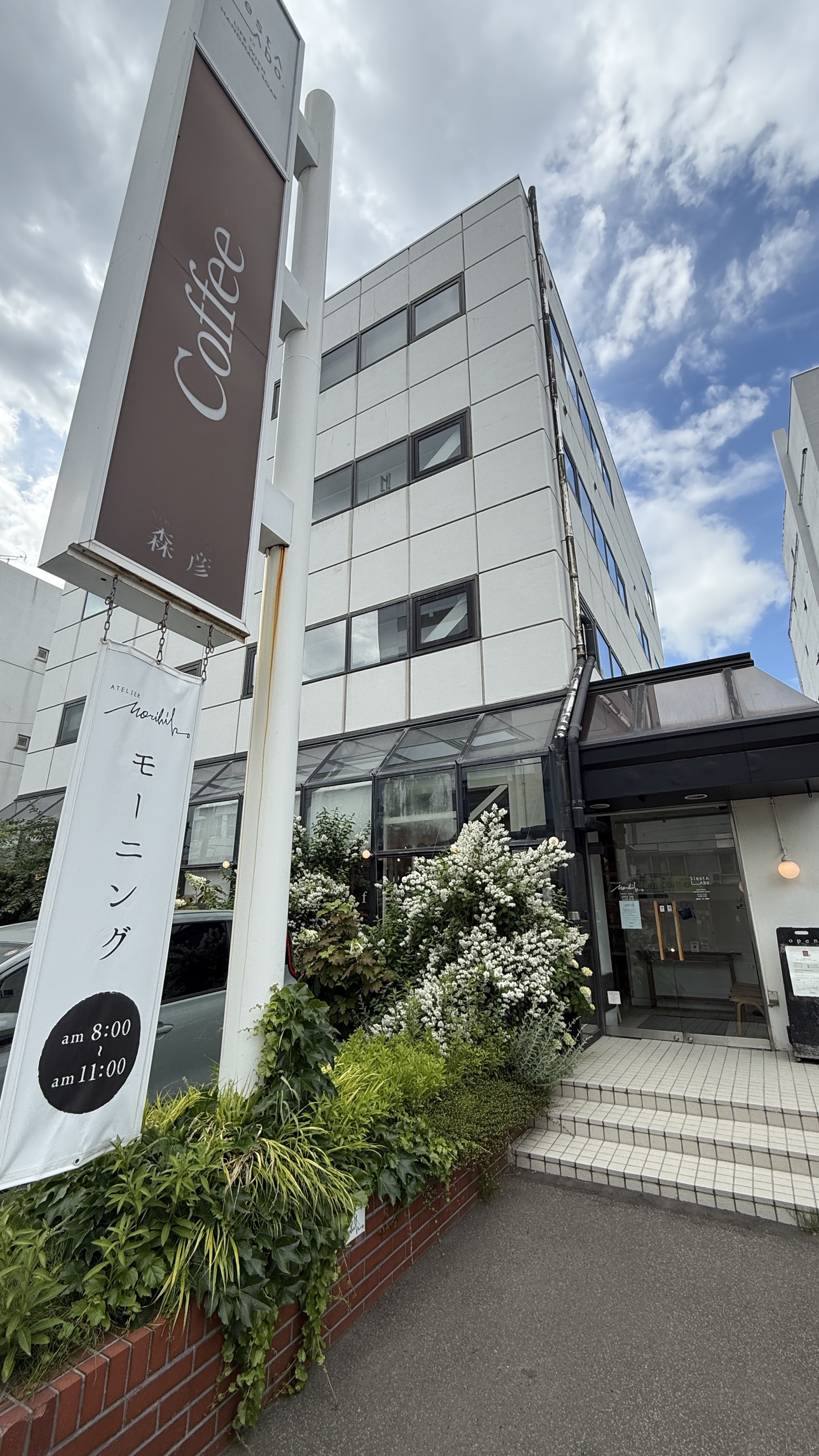 アトリエ森彦の店頭。「Coffee 森彦」の縦看板とモーニング案内のバナー、白い建物の落ち着いた入口