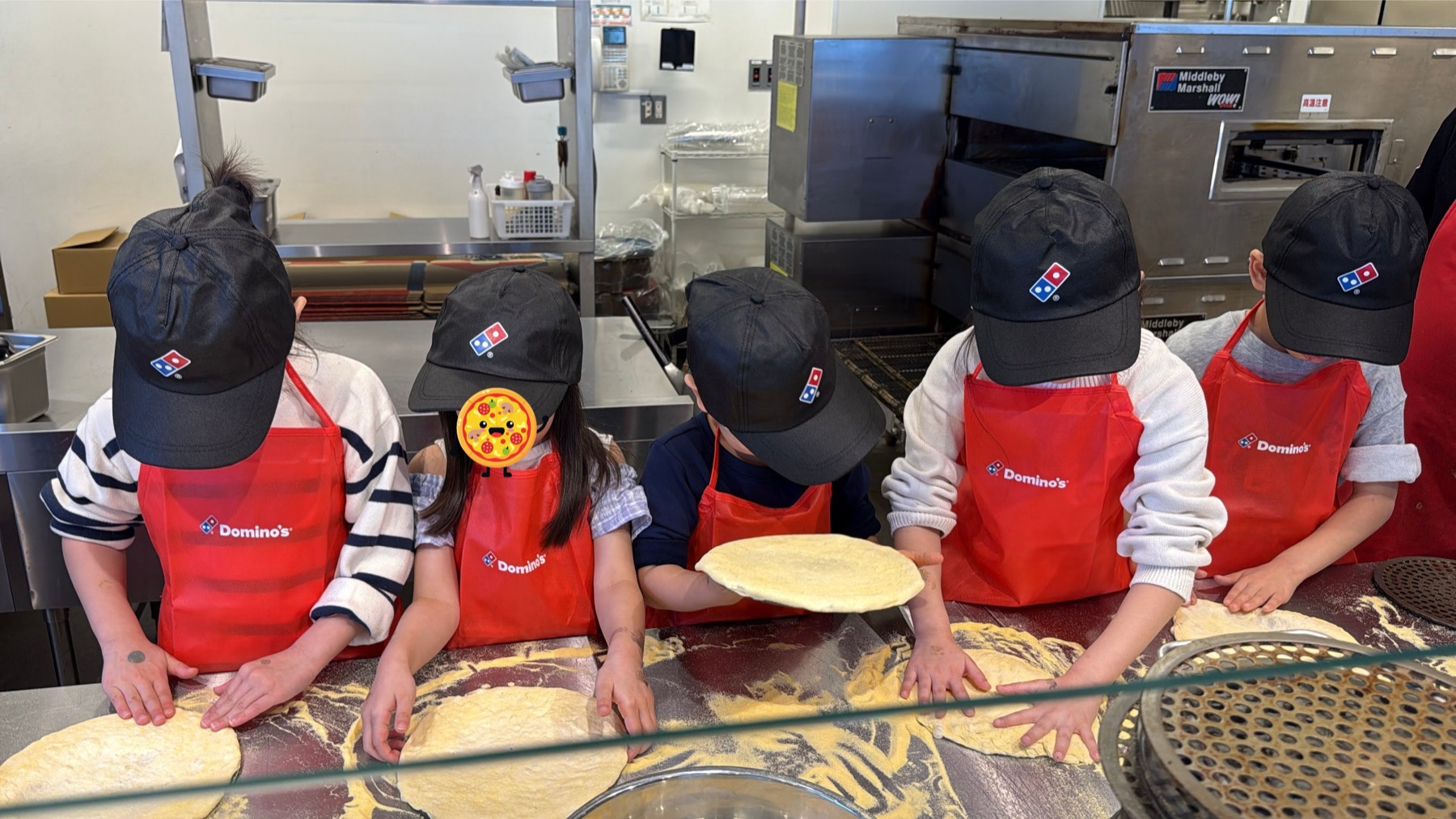 ドミノピザのキャップとエプロンを身につけて、ピザ生地をのばす5人の子どもたち。本物の厨房で本物の生地と向き合っている
