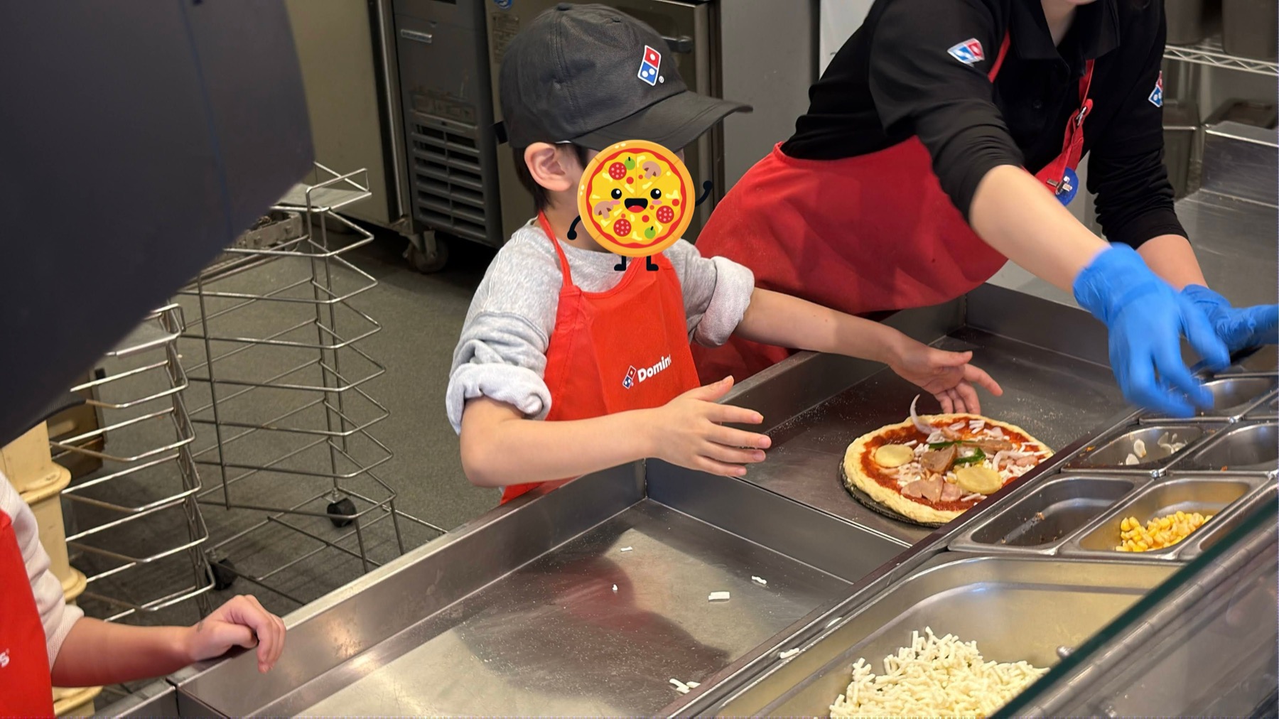 ピザ生地にトッピングをのせている子ども。手前にはピザソースがかかった生地、横にはコーンやチーズの大きなバットが並んでいる