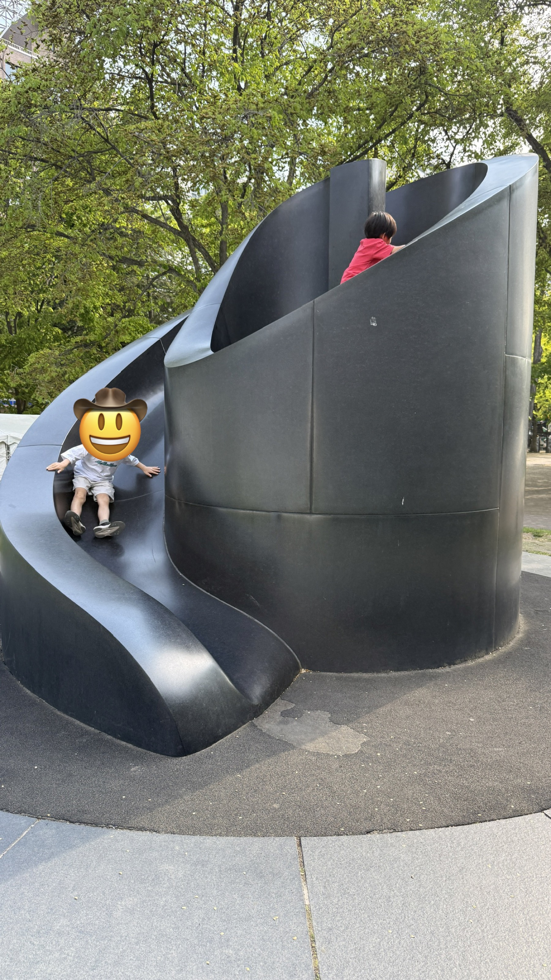 大通公園の中にある黒い大きな滑り台型遊具。次男が滑り台の途中で座り、長男はその上の方へよじ登っている。プライバシー保護のため次男の顔は絵文字で隠してあります