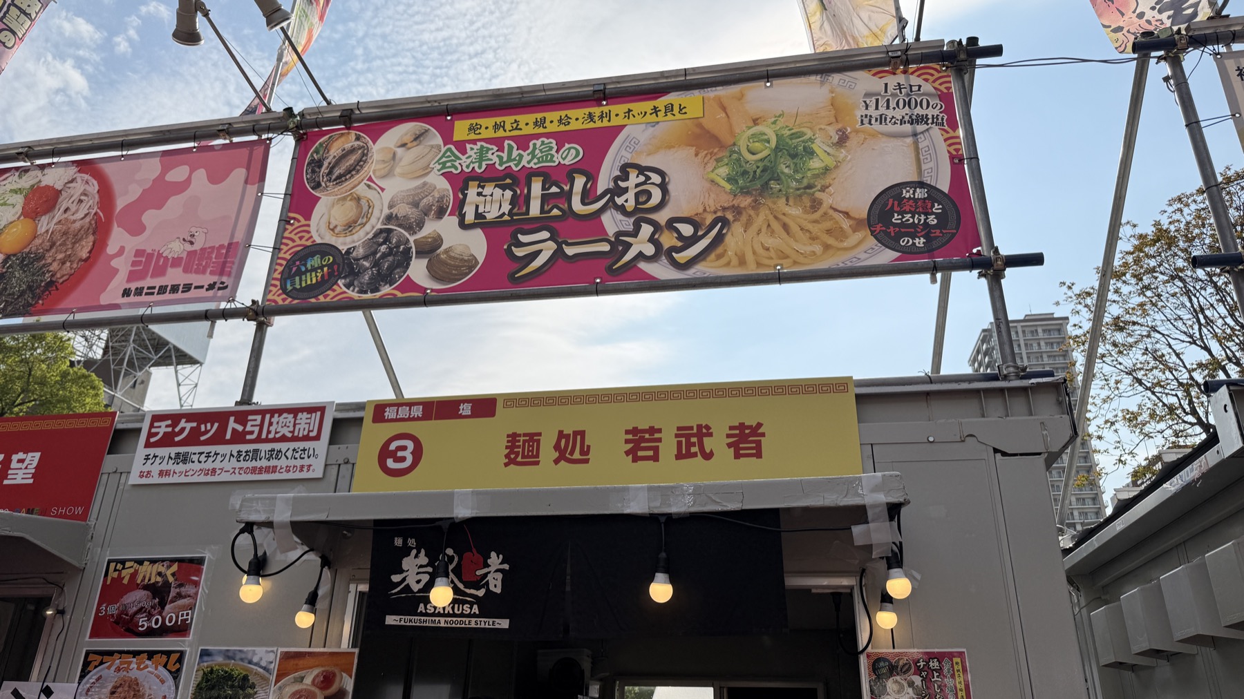 札幌ラーメンショー2026の屋台。「会津山塩 極上しおラーメン」と書かれた大きな看板、奥に「麺処 若武者（ASAKUSA・FUKUSHIMA NOODLE STYLE）」の店構え