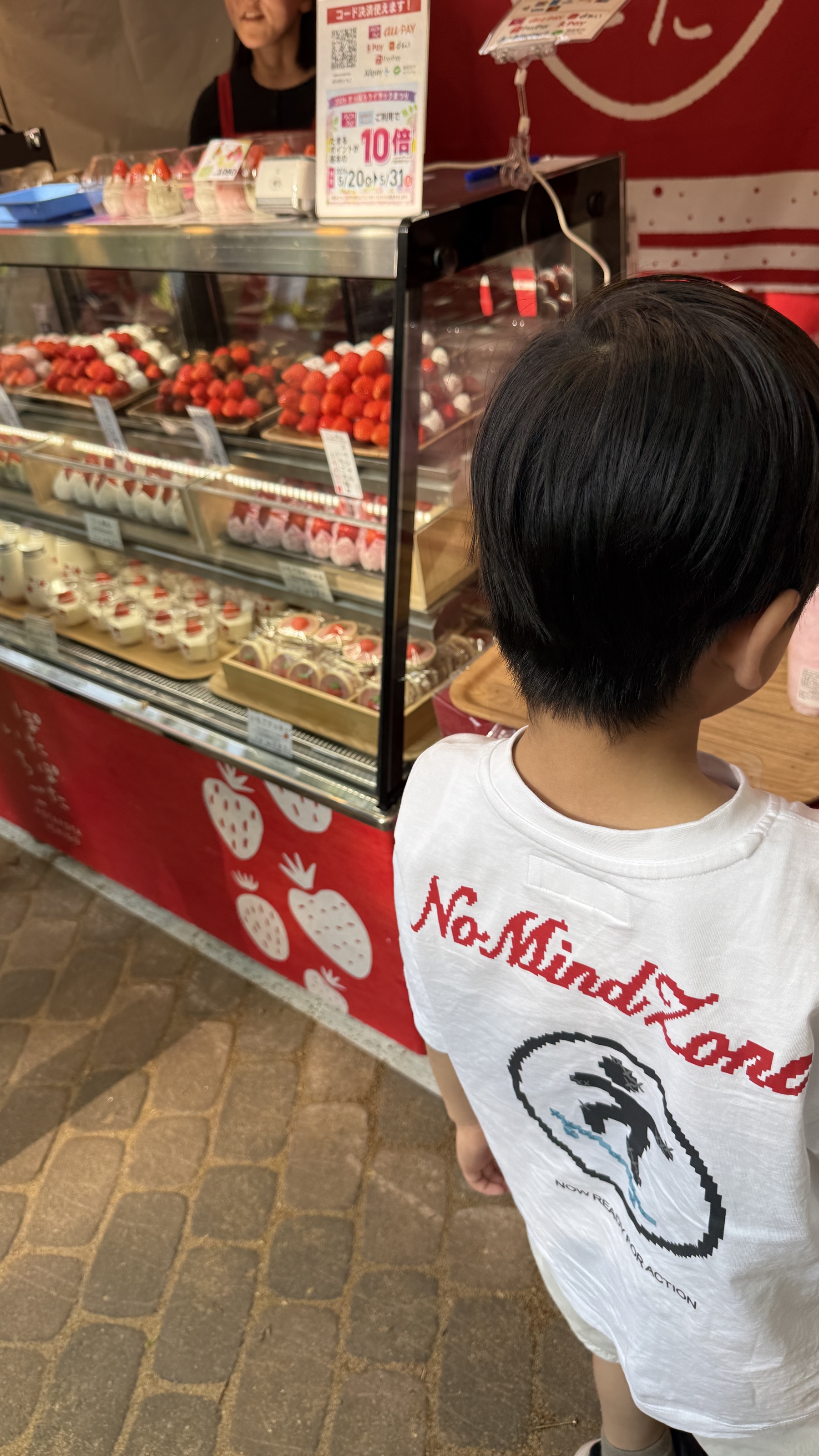 ぽたぽたいちごのショーケースを真剣に見つめる長男の後ろ姿。白Tシャツに、いちごのデコレーションが施された赤いお店の看板を背に立っている