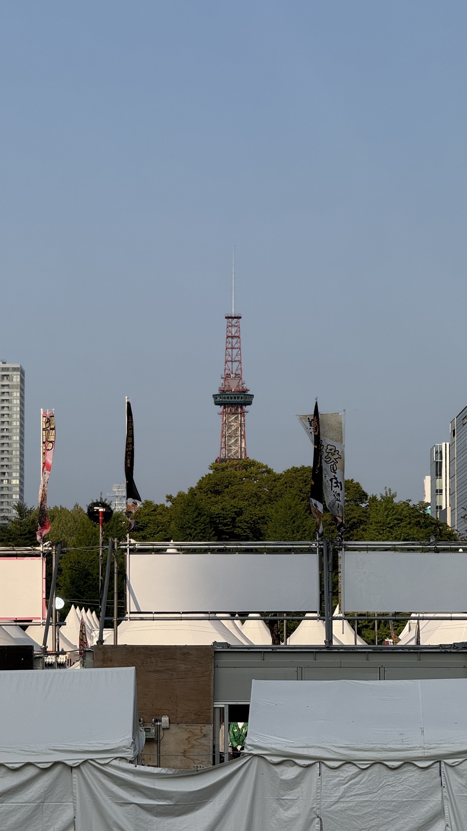 札幌の象徴・さっぽろテレビ塔と、ライラックまつり会場の屋台幟。青空の下、テレビ塔の赤い鉄塔と、屋台の黒い幟、白いテントが並ぶ