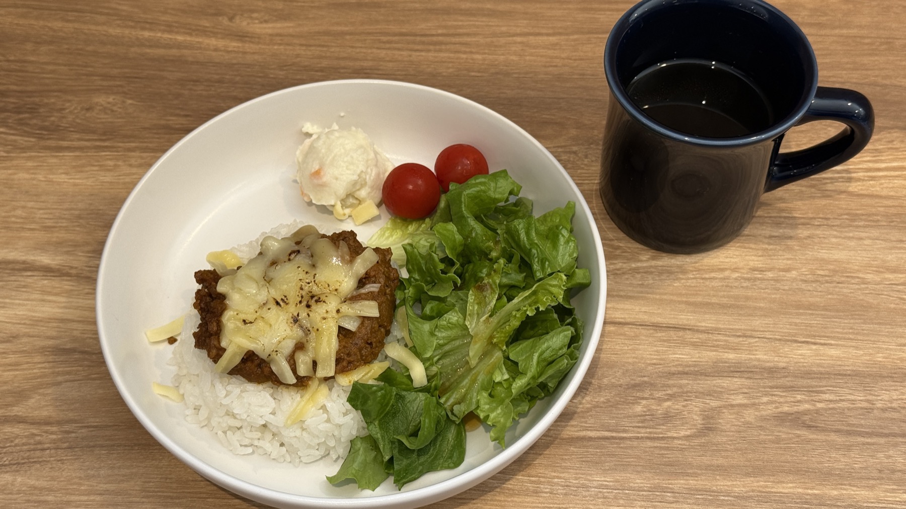 Cafe de raiseのチーズキーマカレー。白いお皿に盛られたごはんとキーマカレーの上にチーズがかかっていて、サラダとポテトサラダ、ミニトマトが添えられている。横に黒いマグカップ