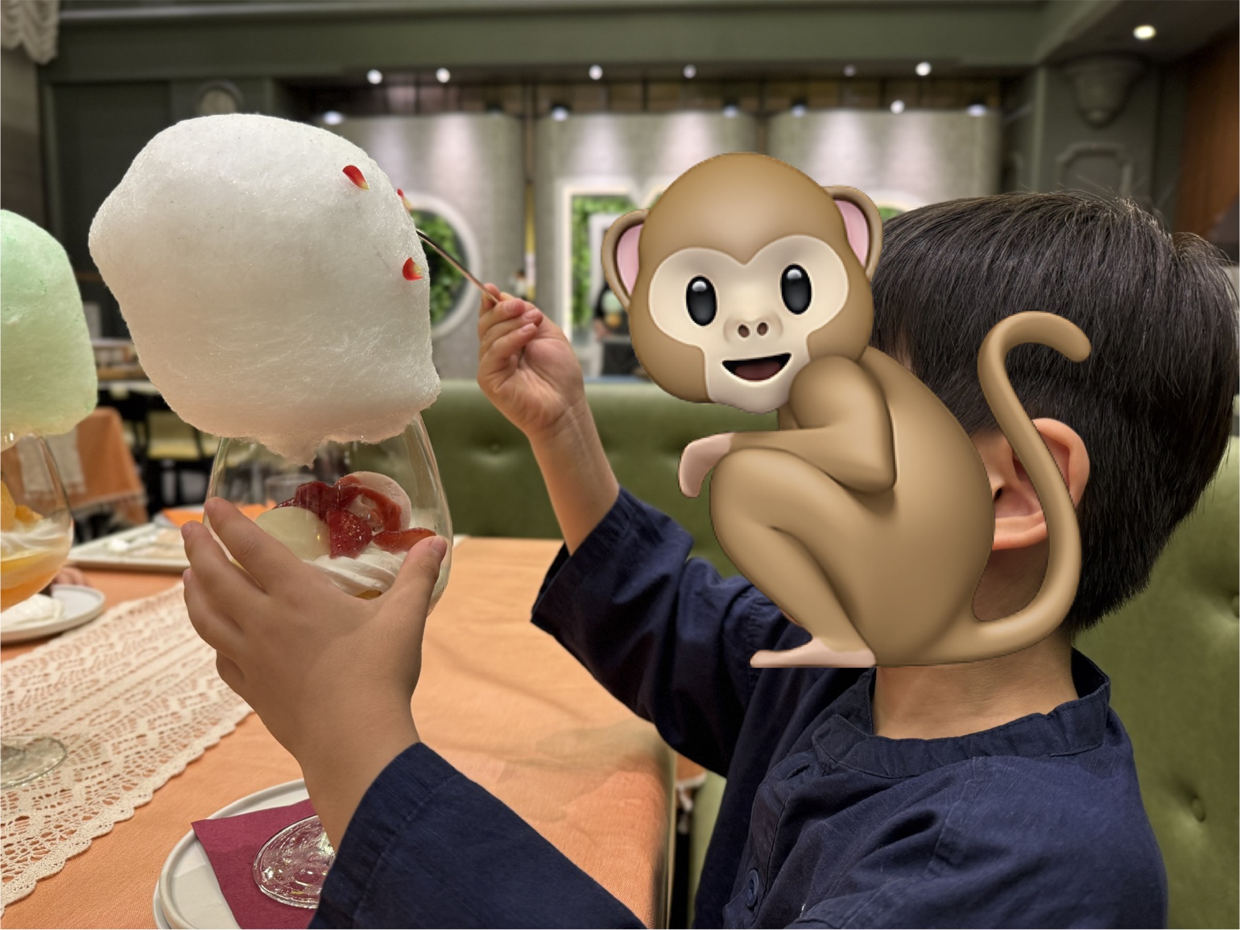 子どもがわたあめパフェを手に持って食べている瞬間。白いわたあめのドームに赤いさくらんぼがトッピング、グラスの底にはイチゴやフルーツが入っている。レストランの緑のソファが背景に