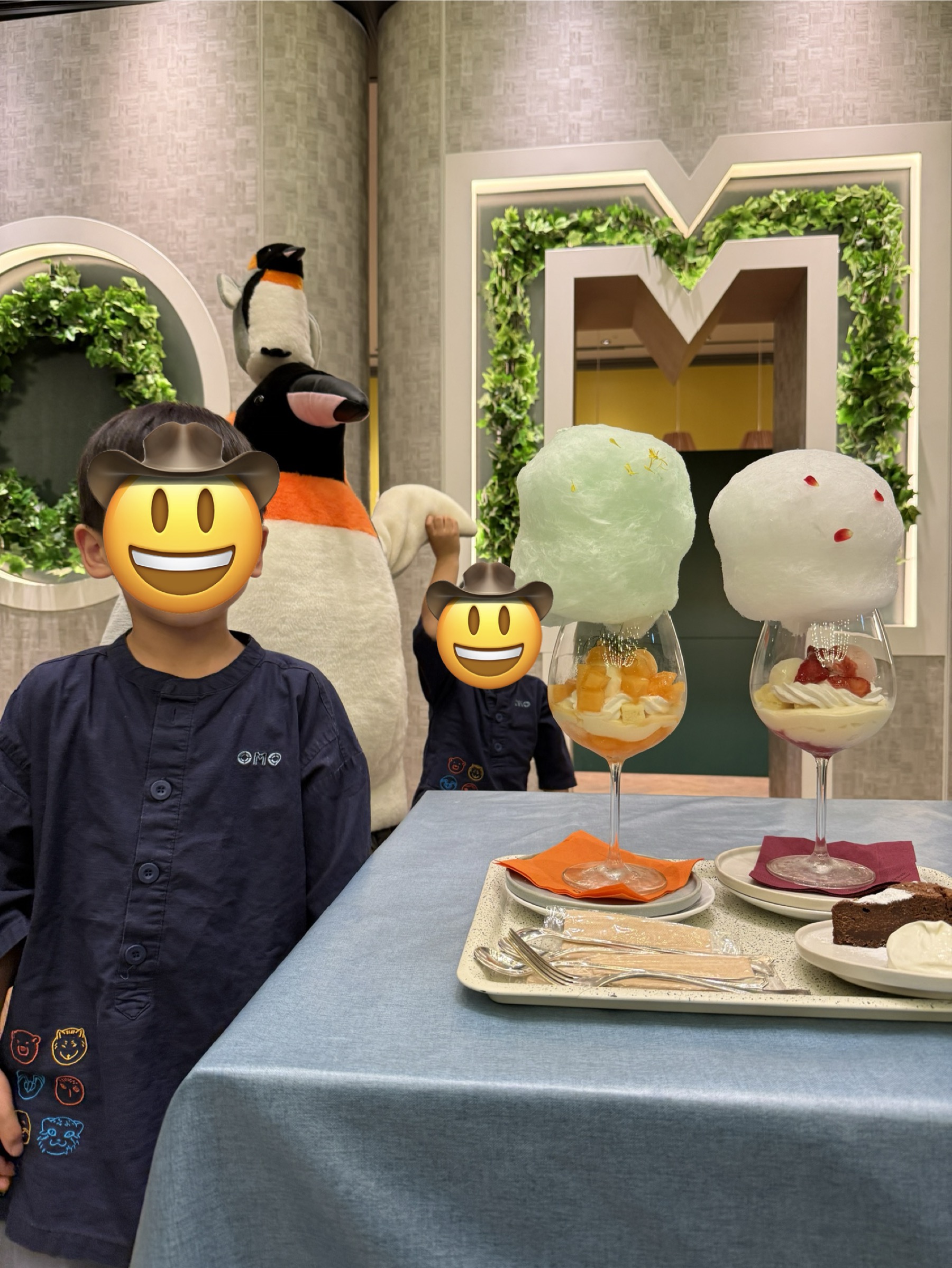 OMOカフェ＆バルの季節限定わたあめパフェ。緑のわたあめが乗ったパフェと白いわたあめが乗ったパフェの2種類が並び、後ろにはホテルのMロゴと旭山動物園をモチーフにしたぬいぐるみ装飾。子どもたちの顔ほどの大きさのわたあめ