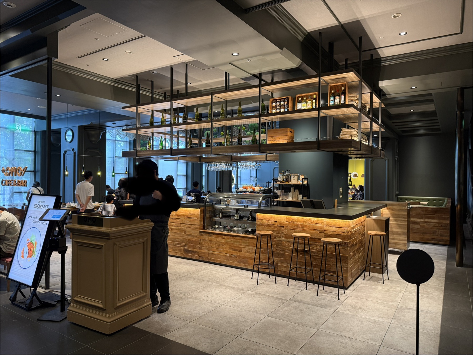 OMOカフェ＆バルの店内。木製のカウンターと天井から吊り下げられた棚のディスプレイ、奥にはバーカウンターの椅子が並ぶ。落ち着いた照明とモダンなインテリア